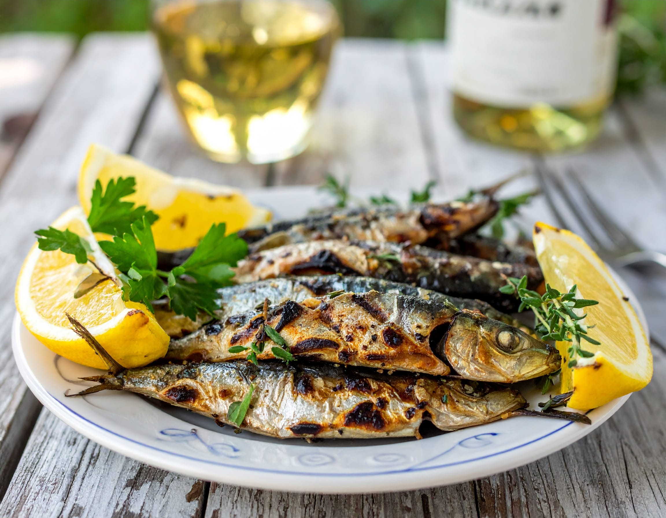 rustic Mediterranean outdoor grill scene with grilled sardines on a plate, garnishe 872840 Grilled sardines served with lemon wedges and fresh herbs on a white plate, ideal for a Mediterranean-style dinner