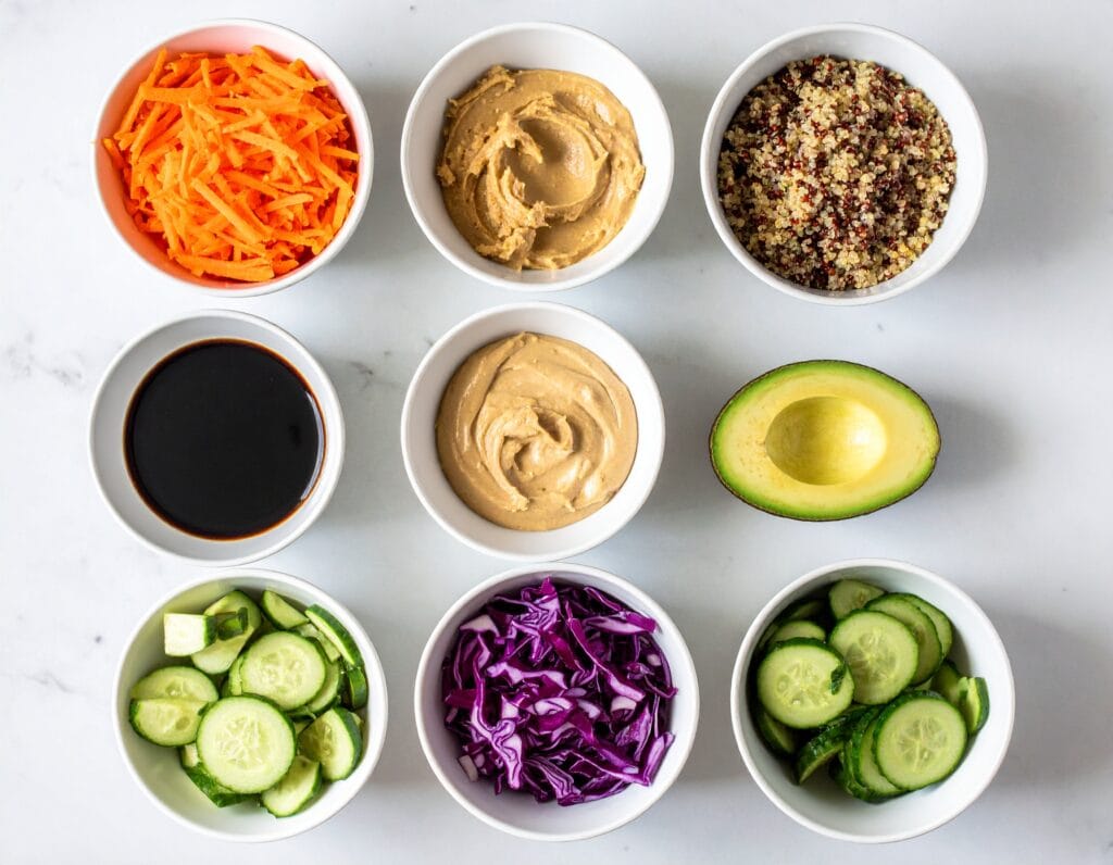 Glass meal prep containers filled with wellness bowl ingredients including rice, vegetables, and peanut carrot slaw on a marble countertop.